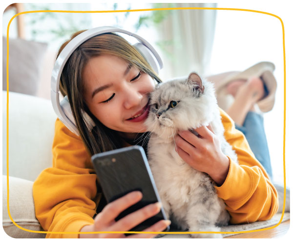 Where pet parents discover brands: real moments, real needs Pet parent lying on a couch, wearing headphones and smiling while holding a smartphone and cuddling a fluffy grey cat.